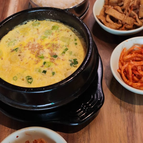 A black bowl of J's Korean Food's steamed egg with green onions and sesame seeds, surrounded by side dishes.