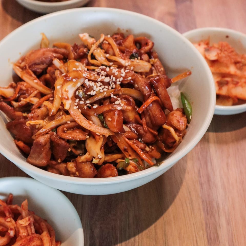 A white bowl of J's Korean Food's spicy stir-fried pork with onions and sesame seeds.