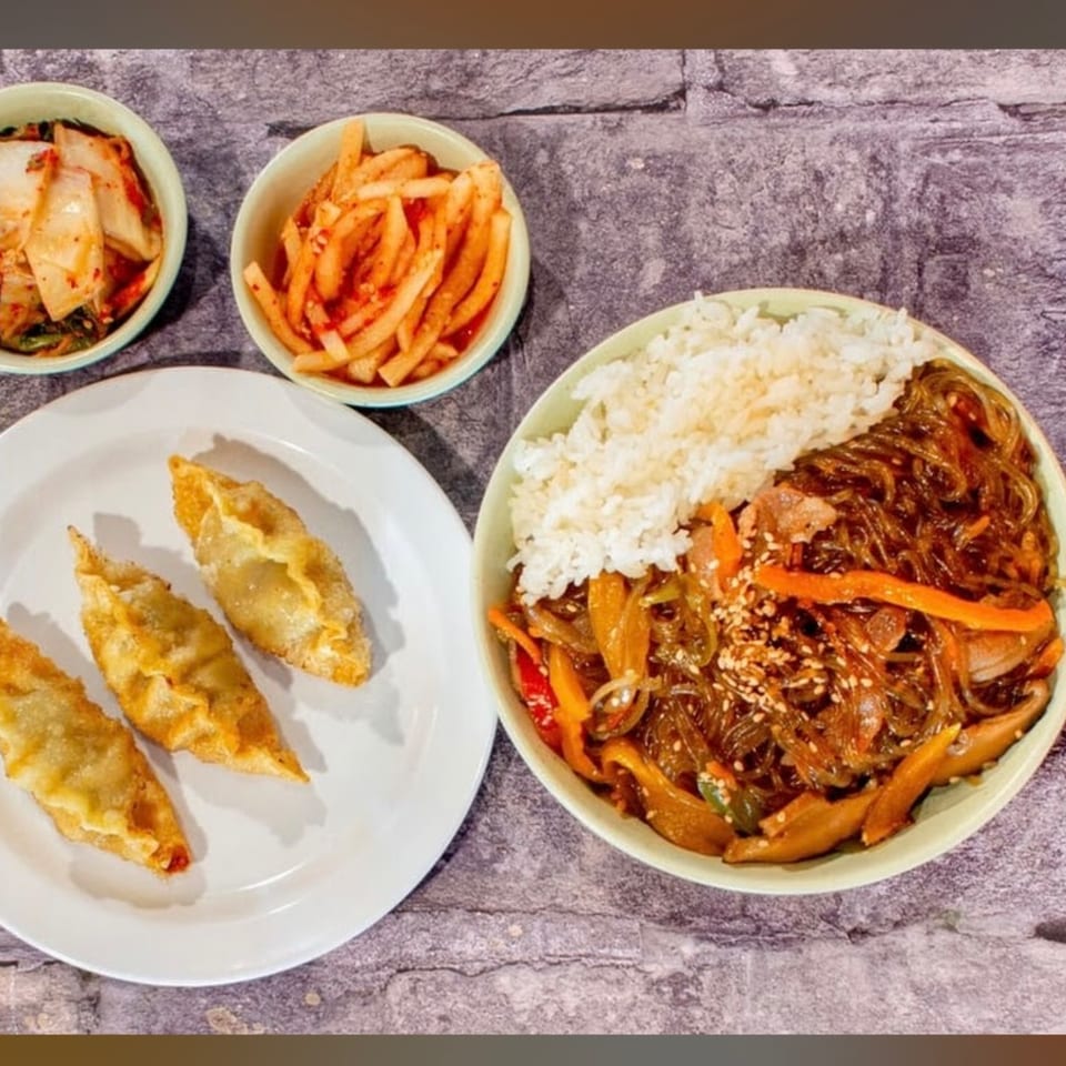 A Korean meal with japchae and rice, fried dumplings, kimchi, and pickled radish on a stone background.