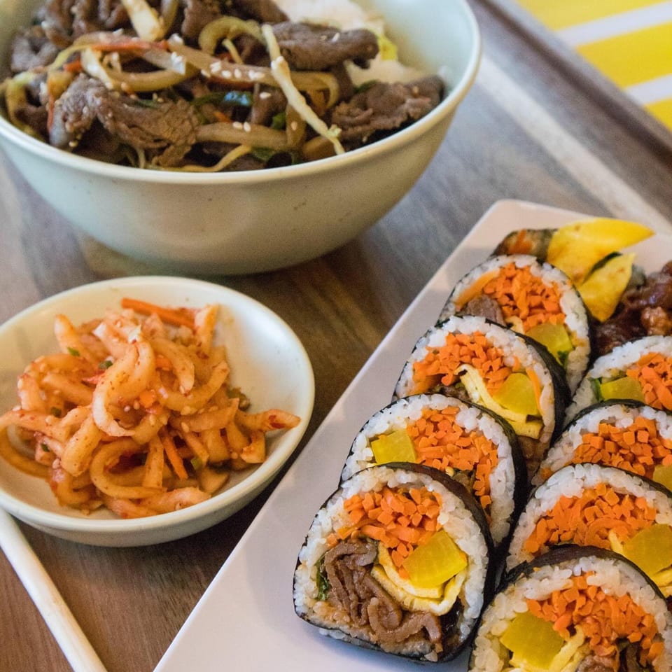 A Korean meal with bulgogi rice bowl, kimchi, and gimbap rolls with beef, egg, and carrots.
